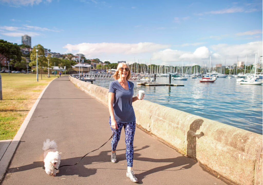 Woman enjoying the health benefits of walking her dog with coffee in hand along the water