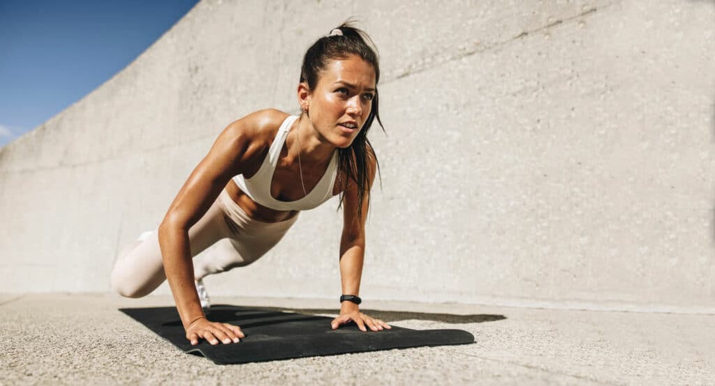 Woman in white activewear doing mat pilates exercise in front of textured concrete wall